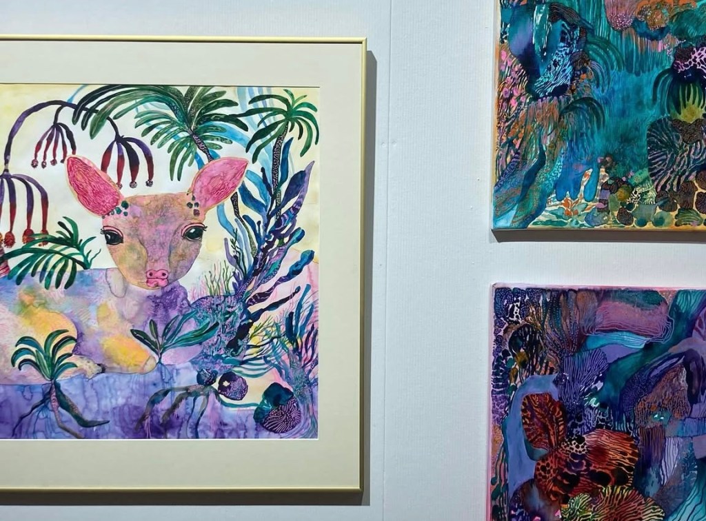 A colorful painting featuring a deer with pink ears, surrounded by vibrant plants and abstract shapes, displayed alongside two other detailed artworks in shades of blue and purple.